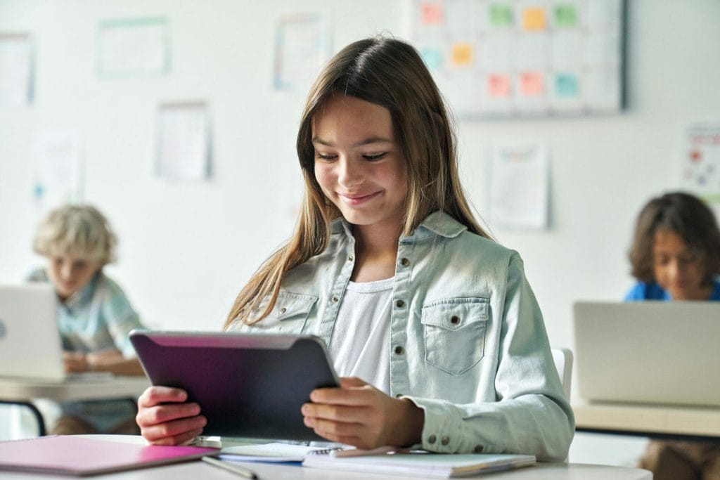 Glücklich lächelnde Schülerin, die eine Aufgabe auf einem Tablet-Computer im Klassenzimmer liest.