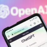 Smartphone zeigt OpenAI-Webseite mit ChatGPT-Interface auf einem Bildschirm, unscharfes OpenAI-Logo im Hintergrund.
