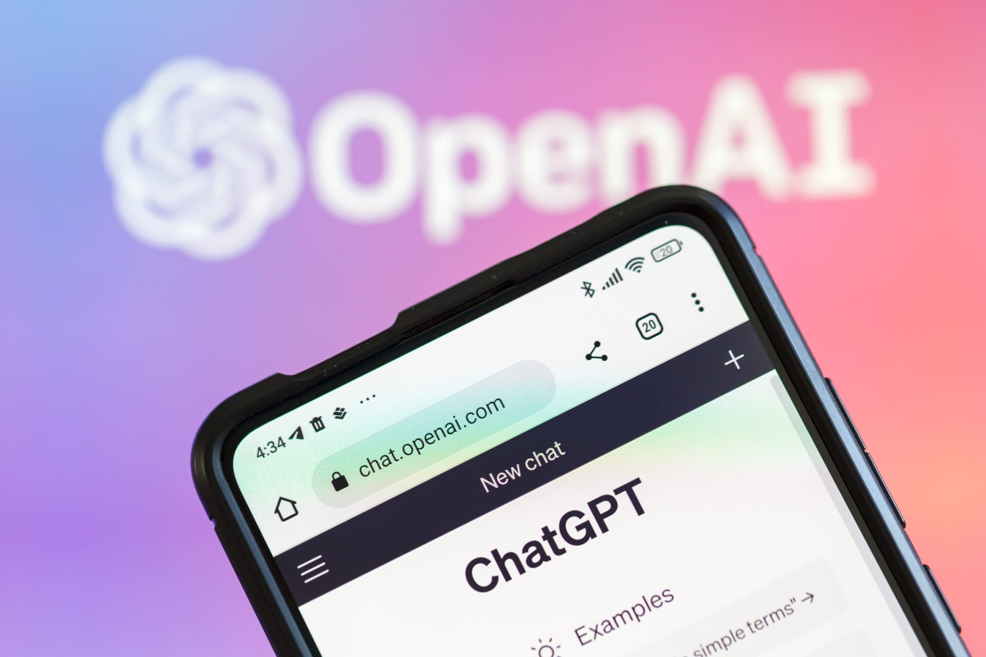 Smartphone zeigt OpenAI-Webseite mit ChatGPT-Interface auf einem Bildschirm, unscharfes OpenAI-Logo im Hintergrund.