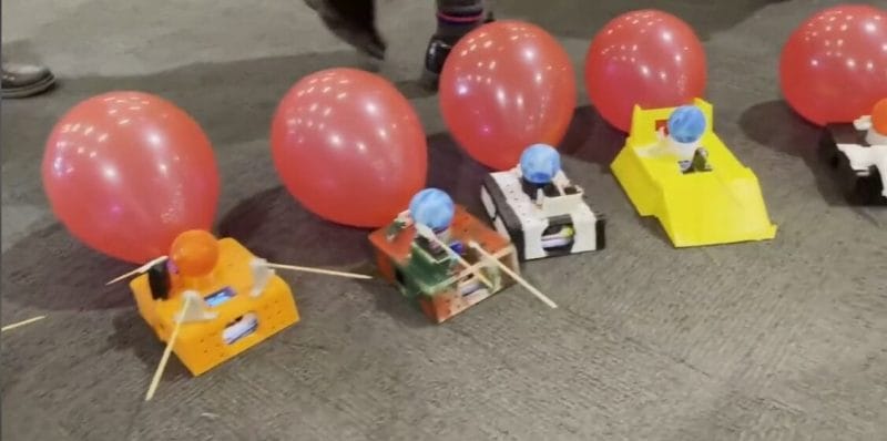 Selbstgebaute Roboterfahrzeuge mit Luftballons in einer Ausstellungshalle, Menschen im Hintergrund.
