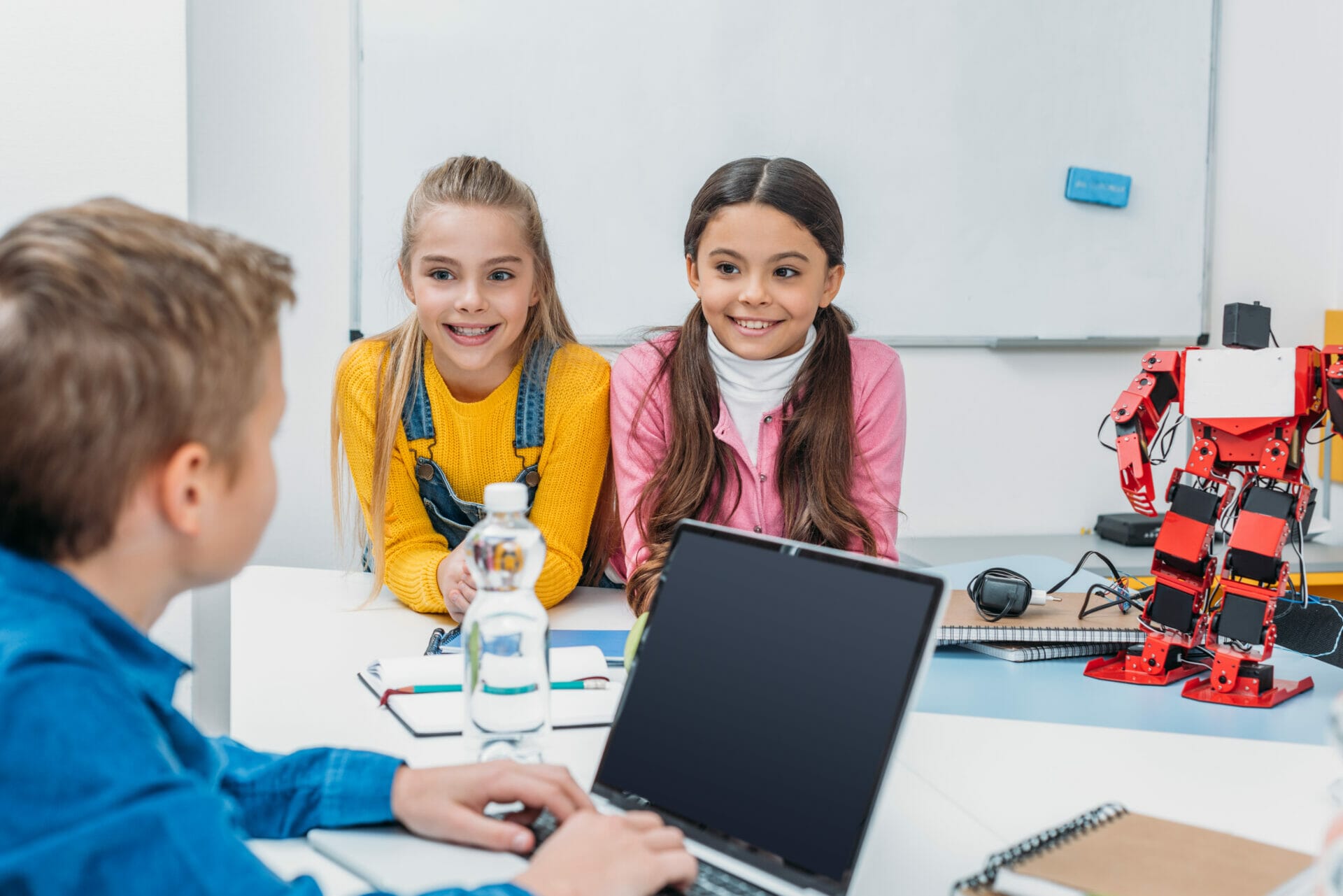 Ein Junge sitzt an einem Laptop am Tisch. Zwei Mädchen lachen ihn an. Es befindet sich ein Roboter, der gerade programmiert wird, auf dem Tisch.