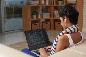 Eine kurzhaarige Frau von hinten gesehen, die an einem Laptop arbeitet, auf dessen Bildschirm Code zu sehen ist, in einem modernen Wohnzimmer mit Bücherregalen und Körben im Hintergrund.