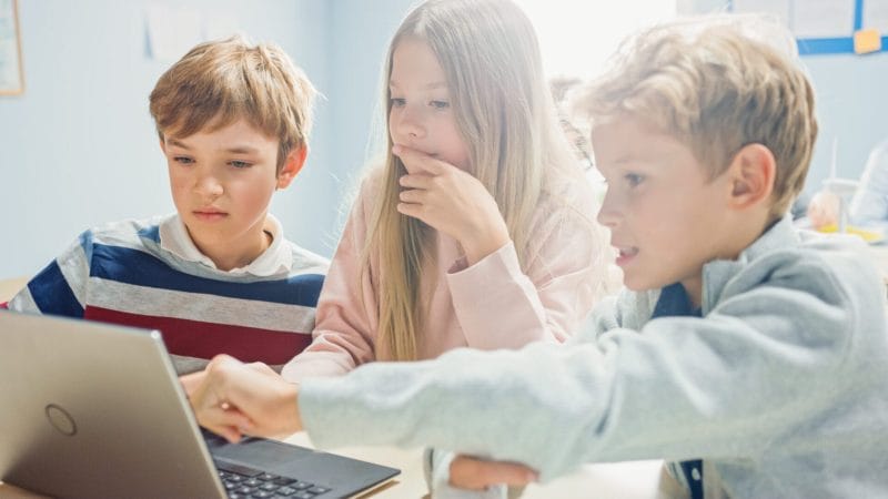 Drei Kinder, zwei Jungen und ein Mädchen, betrachten konzentriert einen Laptop-Bildschirm. Das Mädchen hält ihre Hand vor den Mund, ein Junge zeigt auf den Bildschirm. Sie sitzen in einem hellen Klassenzimmer.