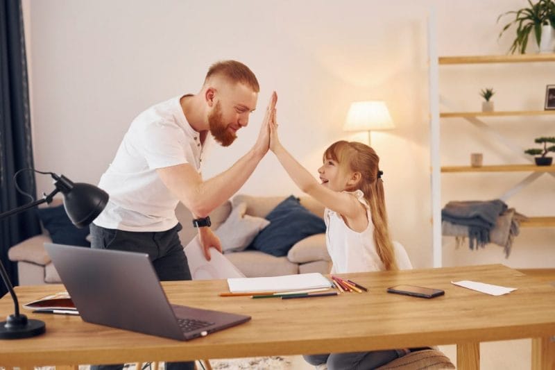 Mann und kleines Mädchen geben sich in einem gemütlichen Wohnzimmer mit einem Schreibtisch und einem Laptop ein High-Five.