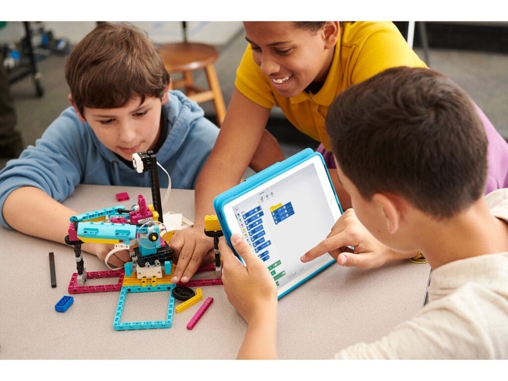 Drei Kinder sitzen um einen Tisch und arbeiten an einem Robotikprojekt mit Legosteinen. Ein Kind hält ein Tablet in der Hand und programmiert möglicherweise, während die anderen beim Bauen helfen.