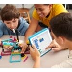 Drei Kinder sitzen um einen Tisch und arbeiten an einem Robotikprojekt mit Legosteinen. Ein Kind hält ein Tablet in der Hand und programmiert möglicherweise, während die anderen beim Bauen helfen.