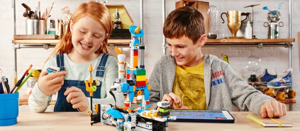 Zwei Kinder sitzen an einem Tisch und spielen mit bunten LEGO-Steinen und einem Tablet. Sie lächeln und scheinen konzentriert zu sein. Im Hintergrund sind verschiedene Werkzeuge und Materialien zu sehen.