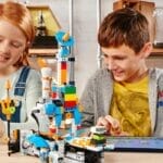 Zwei Kinder sitzen an einem Tisch und spielen mit bunten LEGO-Steinen und einem Tablet. Sie lächeln und scheinen konzentriert zu sein. Im Hintergrund sind verschiedene Werkzeuge und Materialien zu sehen.