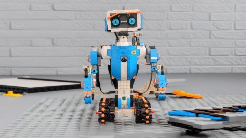 Eine aus Bausteinen zusammengesetzte Lego-Roboterfigur sitzt auf einer grauen Grundplatte. Um sie herum sind verstreut Bausteine verstreut, im Hintergrund ist ein Bausteintrenner zu sehen.