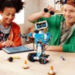 Zwei Kinder bauen und programmieren einen Lego Boost Roboter, den sie aus Bauklötzen gebaut haben. Auf dem Tisch liegen ein Tablet und verschiedene Kleinteile.
