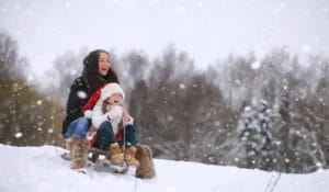 Zwei Menschen genießen einen verschneiten Tag beim Rodeln bergab. Beide sind warm angezogen und Schneeflocken fallen vom bewölkten Himmel.