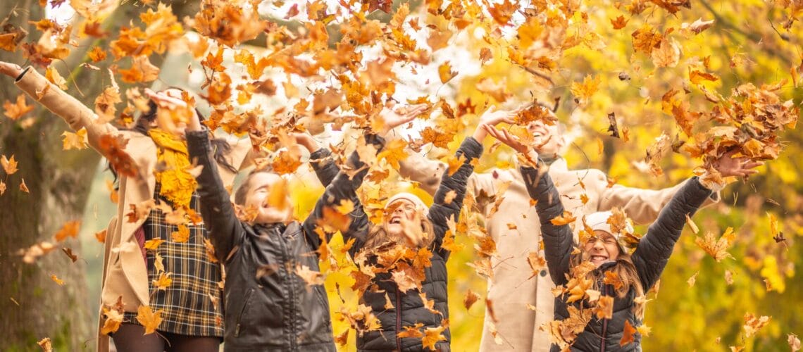 Eine Gruppe von Menschen wirft fröhlich Herbstblätter in die Luft, umgeben von Bäumen mit buntem Herbstlaub.