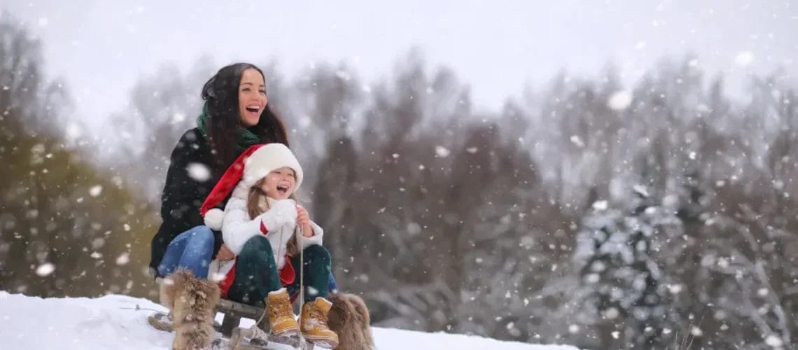 Zwei Menschen genießen einen verschneiten Tag beim Rodeln bergab. Beide sind warm angezogen und Schneeflocken fallen vom bewölkten Himmel.